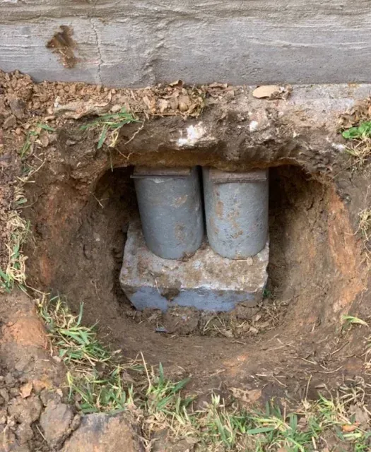 Two gray utility pipes set in a dirt hole beneath a wooden structure.