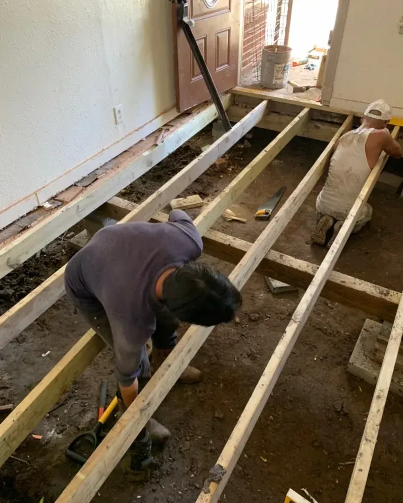 Workers installing floor joists in a room under renovation, with tools and exposed subfloor.