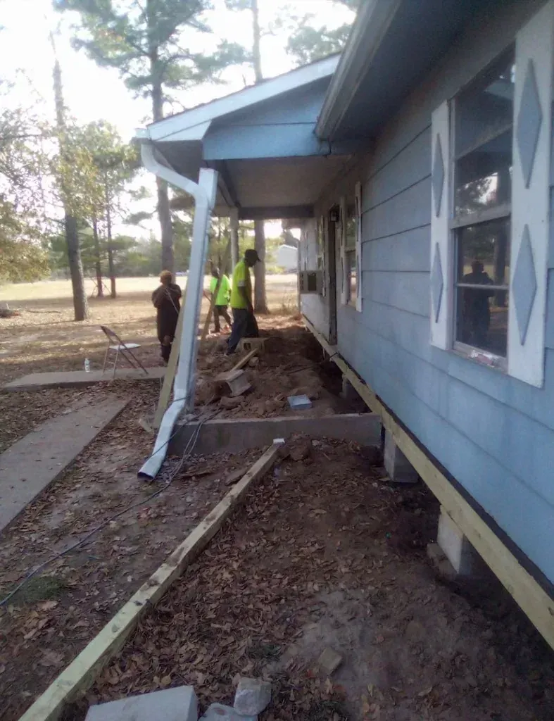 Damaged house exterior with collapsed porch support and debris, two people inspecting the damage