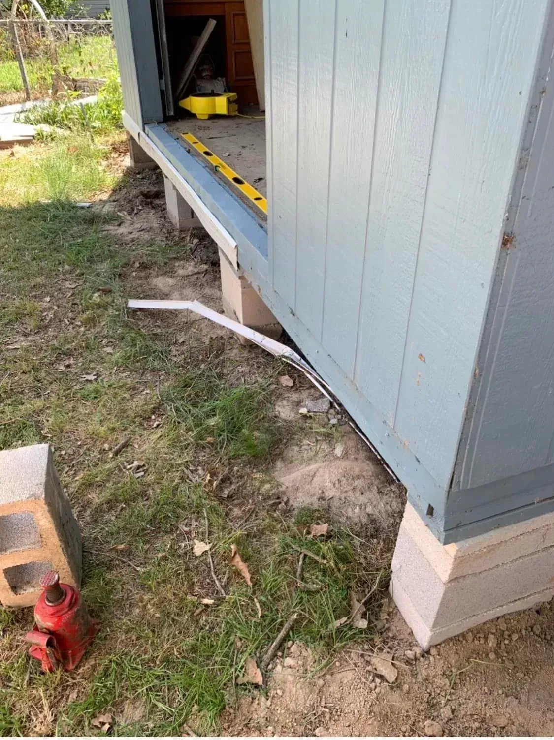 Side of a shed with a raised foundation, yellow workbench inside, and a white pipe lying on the grass.