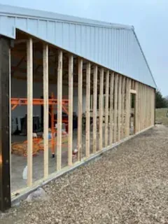 Partially framed building with white metal siding and exposed wooden studs beside a gravel path