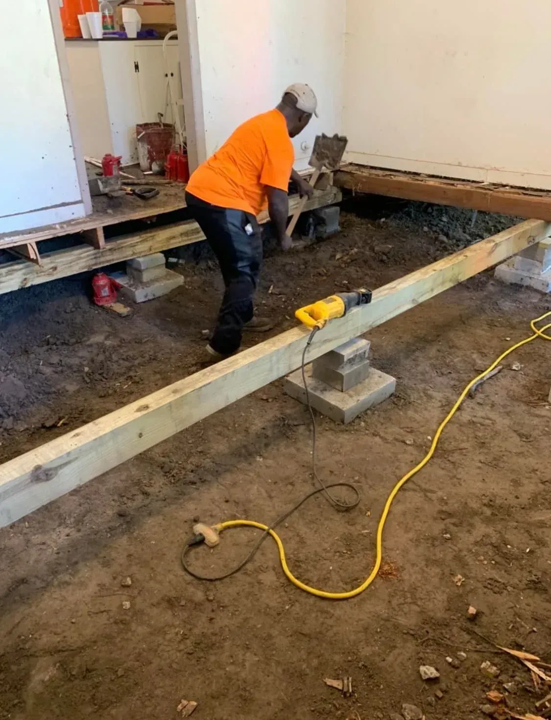 Worker in orange shirt installing a wooden beam inside a construction site with tools and cords on the floor