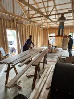 Workers framing a house interior with exposed wooden studs and beams, one standing on a ladder.