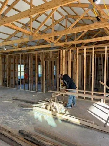 Framed house interior under construction with wooden trusses and a worker handling lumber near the center