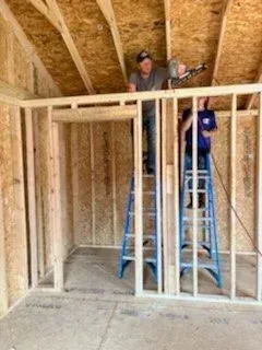 Two workers framing an unfinished interior wall in a house under construction, one on a blue ladder.