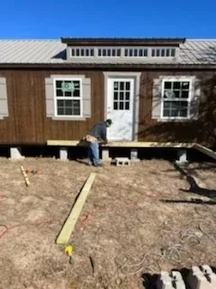 Worker measuring a raised wooden house exterior with a tape on the dirt yard