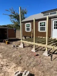 Partially built wooden deck beside a brick house on a dirt yard under a clear blue sky