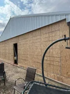Outdoor shed with gray metal roof and plywood siding, seen beside patio chairs and a curved black pipe.