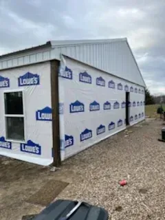 Partially wrapped white shed with Lowe’s logos on exterior, viewed from the corner on a gravel lot.