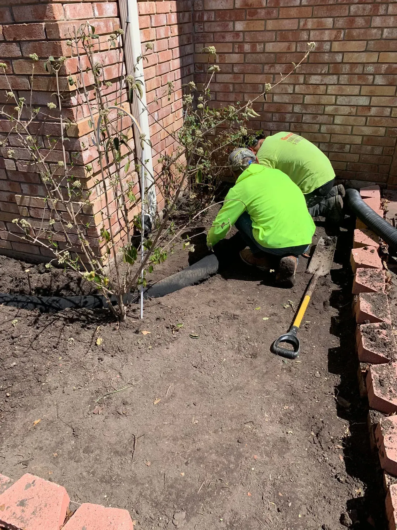 Two workers in neon green shirts clearing debris beside a brick wall and garden bed.