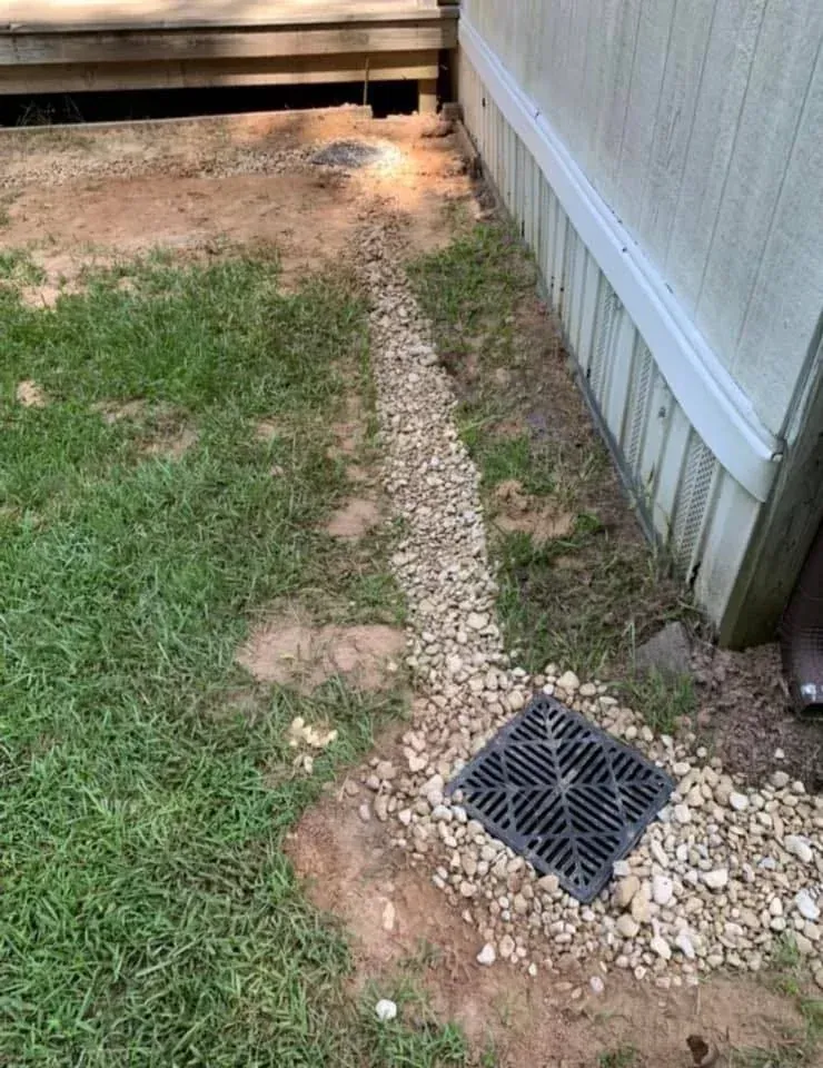 Narrow gravel drainage strip beside a house and fence, with grass and a square drain cover.