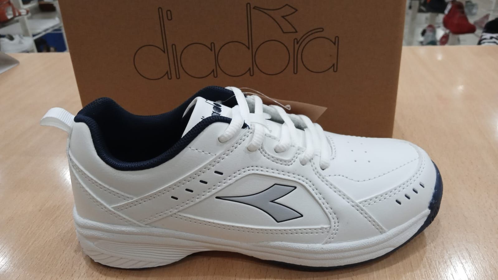 Zapatilla Diadora en color blanco y azul marino con logo y cuadro en el fondo.