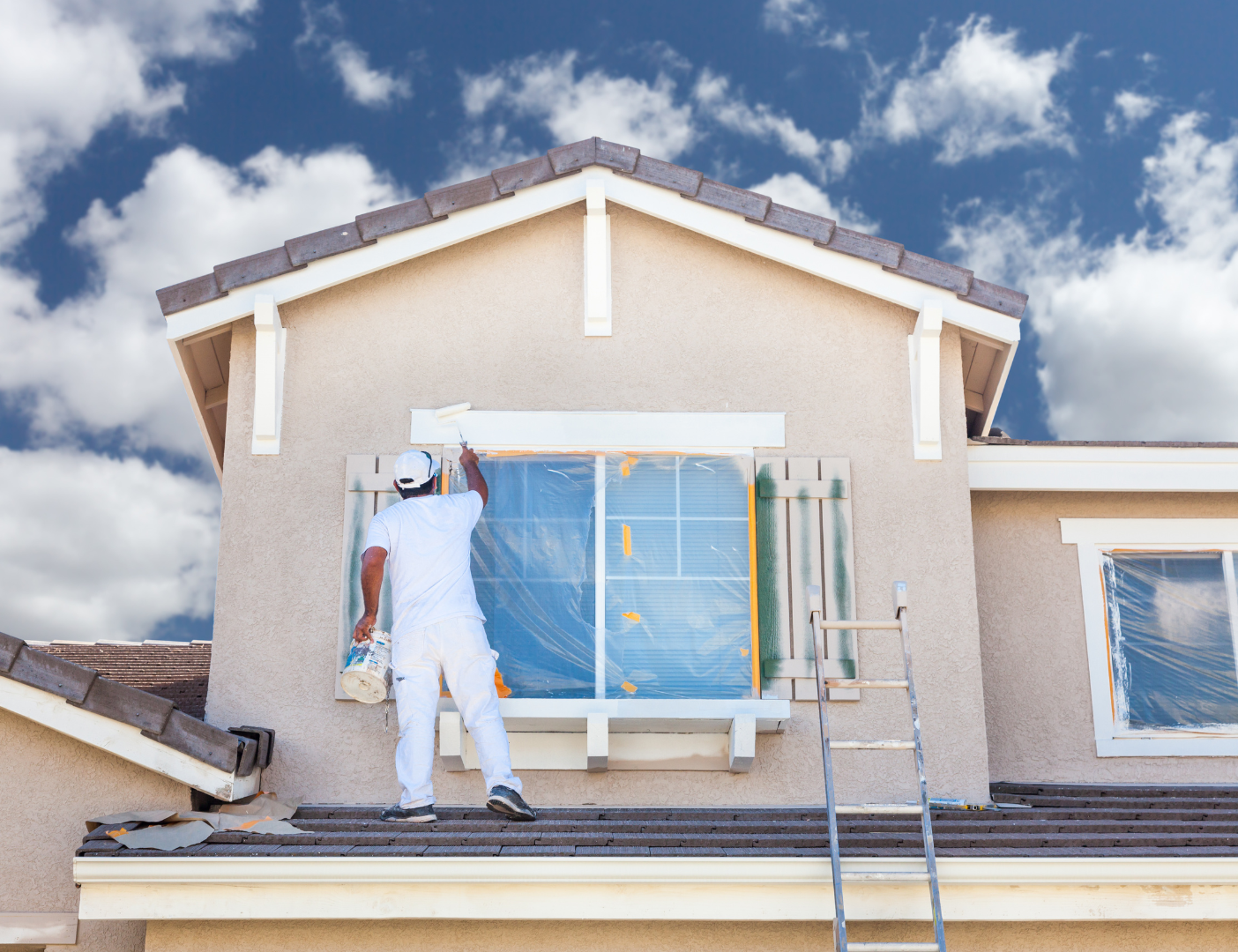 Painter on ladder applying paint to window trim of two-story house, blue sky backdrop.