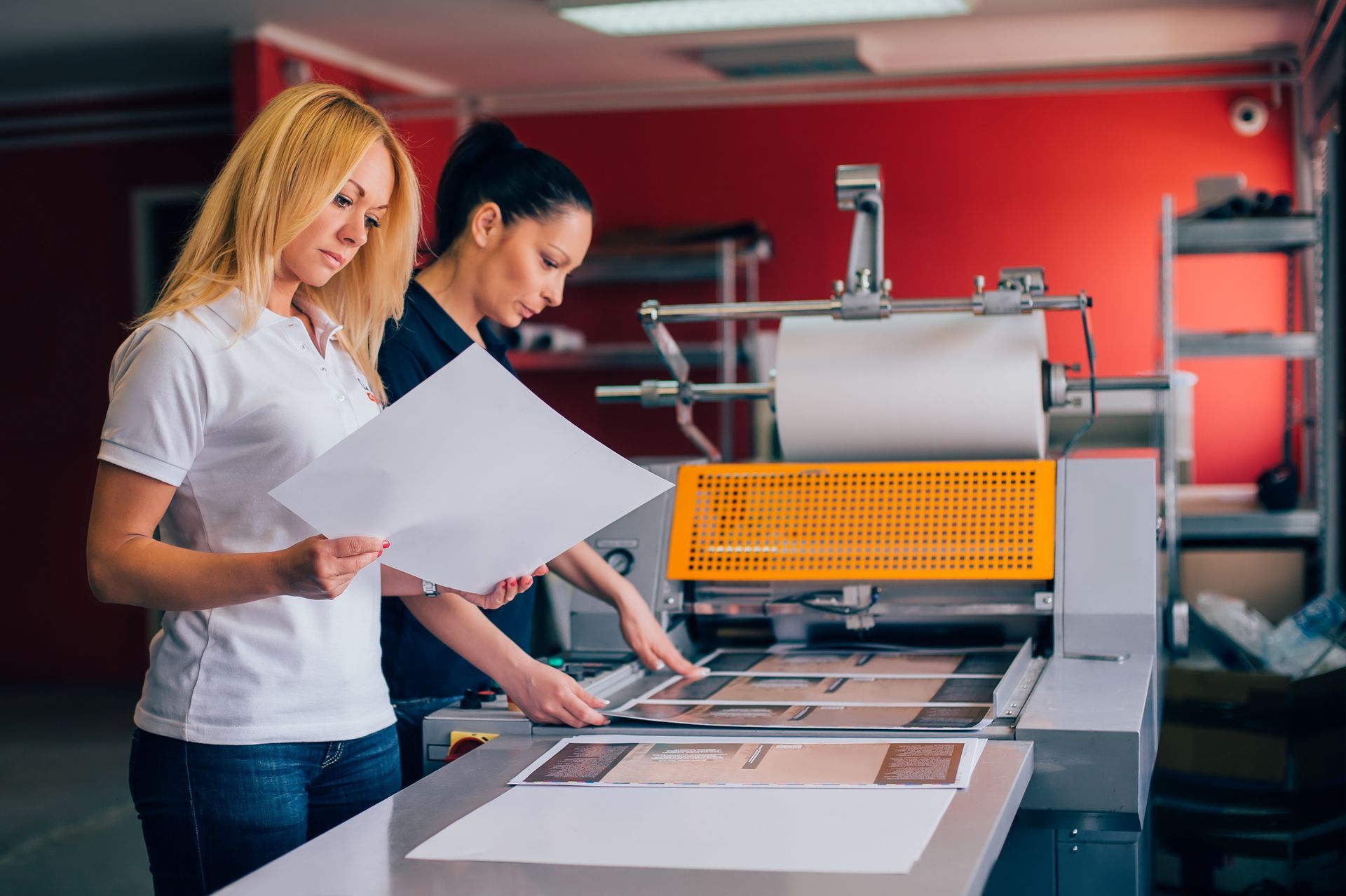 Two people in a print shop inspect a document before placing it into a large paper laminating machine.