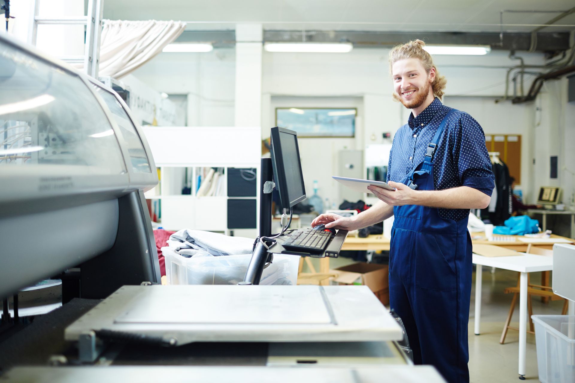 A smiling worker in blue coveralls uses a computer keyboard in a printing or production workspace.