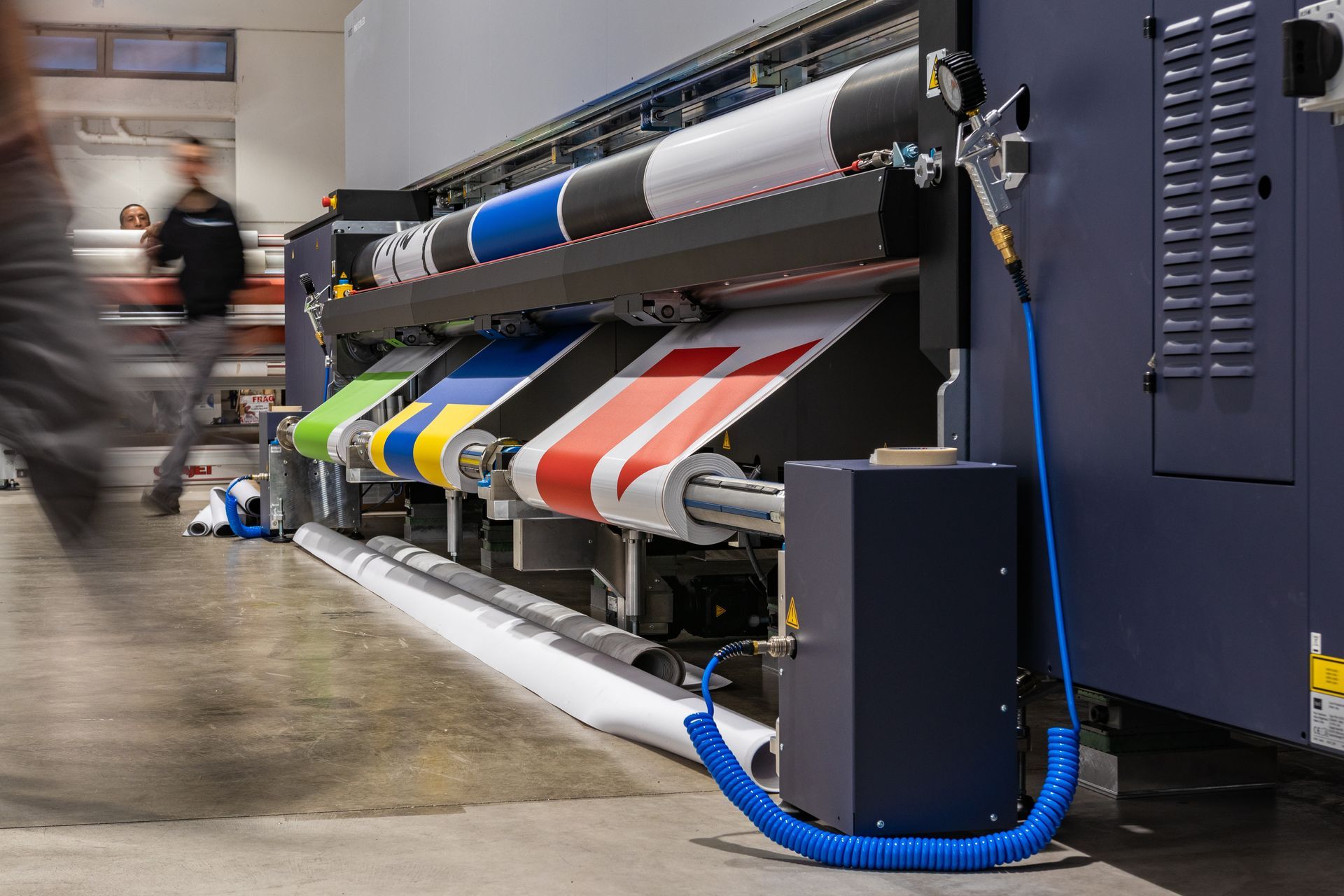 A large industrial printer processes multiple colorful sheets of material in a workshop with a person walking in the background.