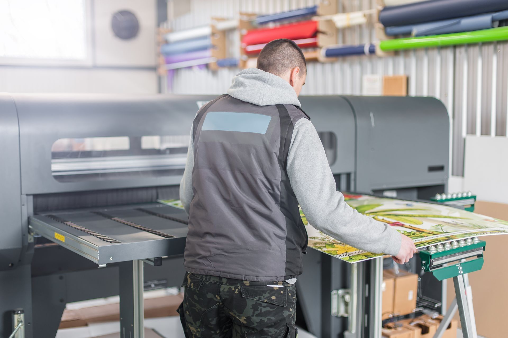 A worker operates a large format printing machine in a warehouse setting, handling a printed sheet of material.