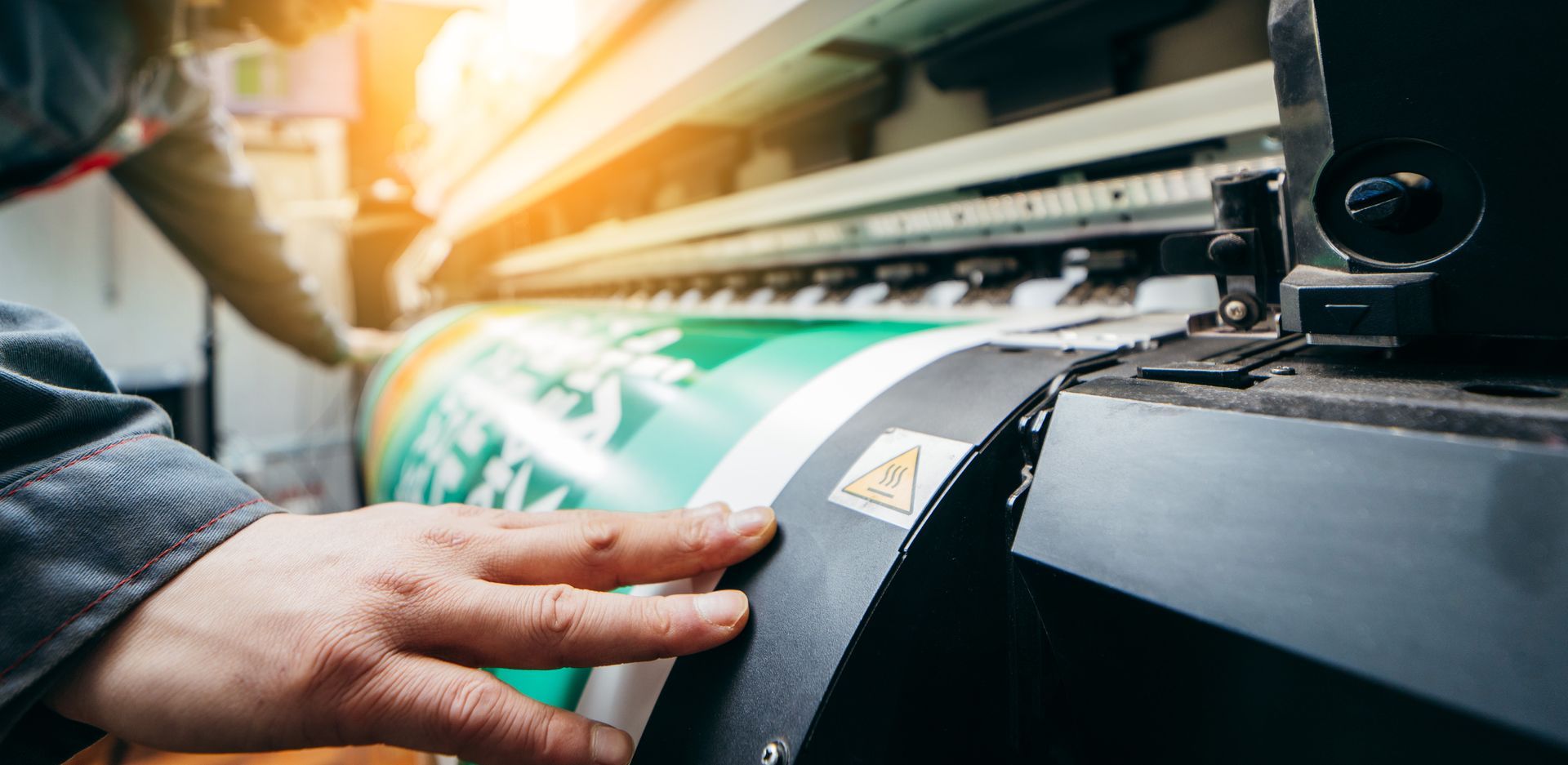 A worker’s hands guide a large green and white printed banner through a professional wide-format printer.