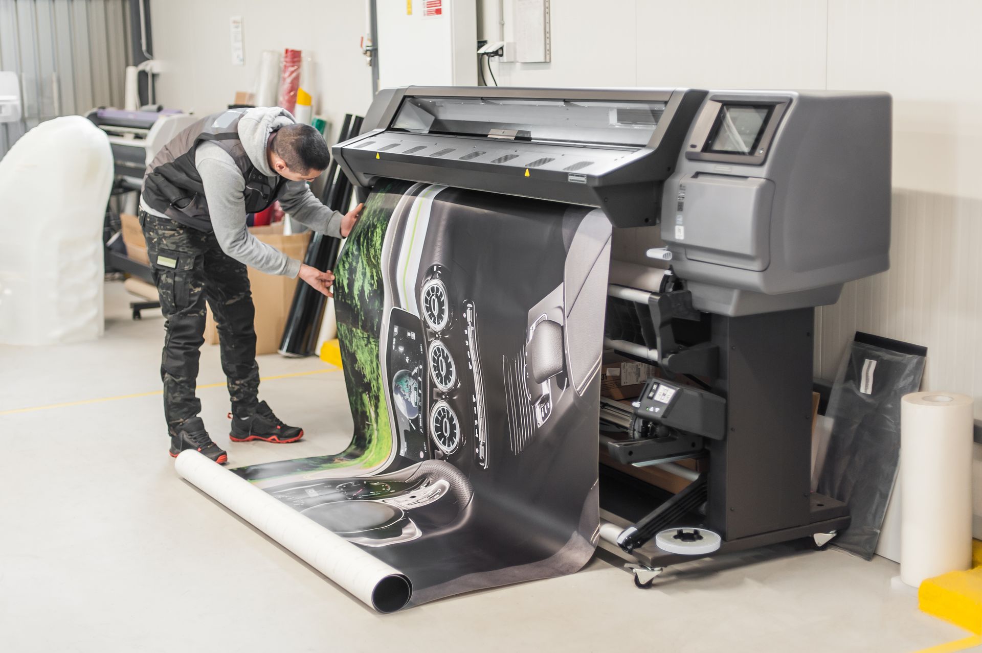 A person in a workshop inspects a large-format printer outputting a graphic display on a roll of vinyl media.