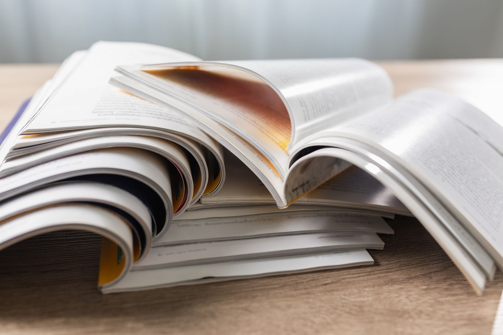 Several open books and magazines are stacked on a wooden surface, their pages fanned out.
