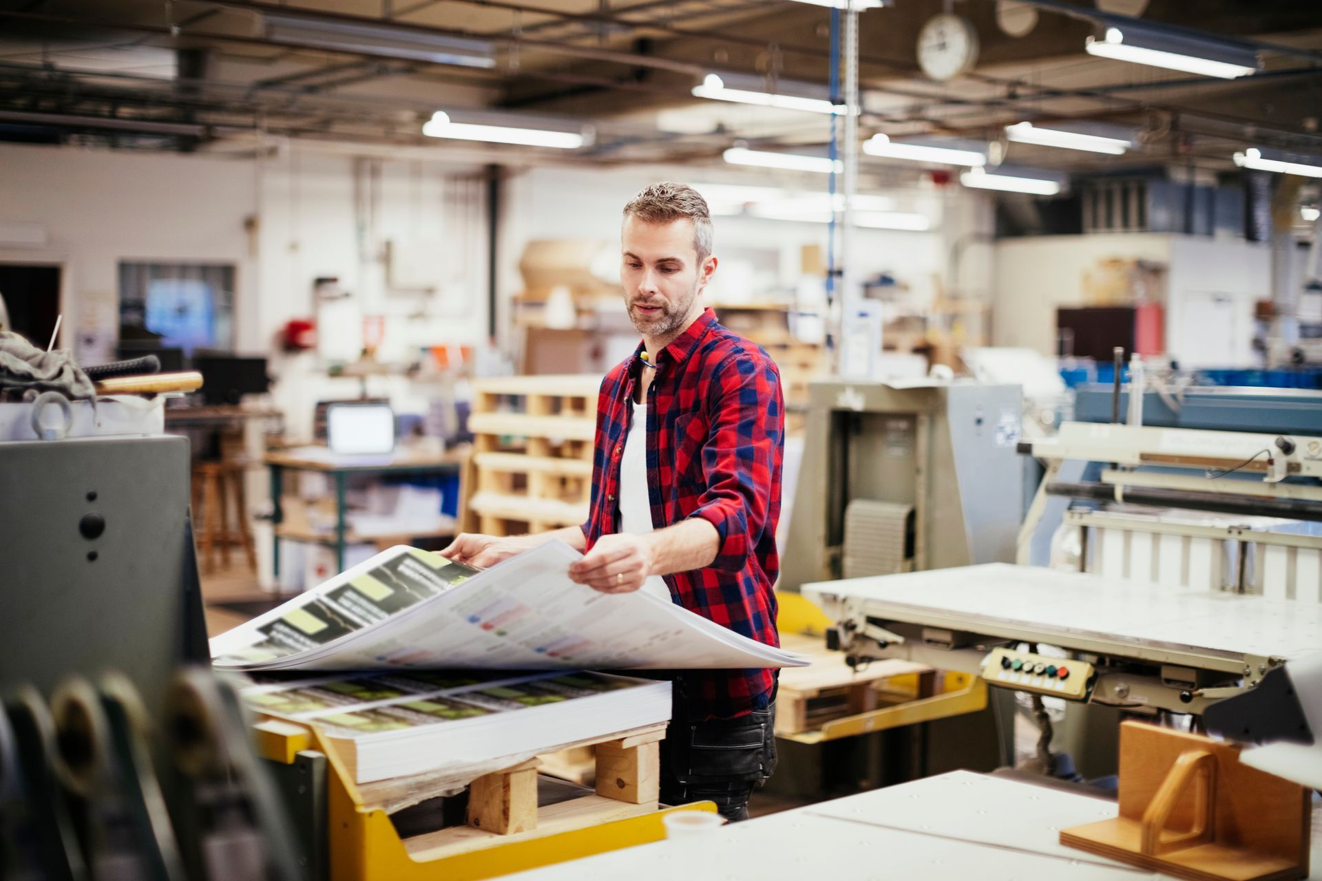 A person in a red plaid shirt inspects a stack of printed materials in a bright, industrial printing shop.