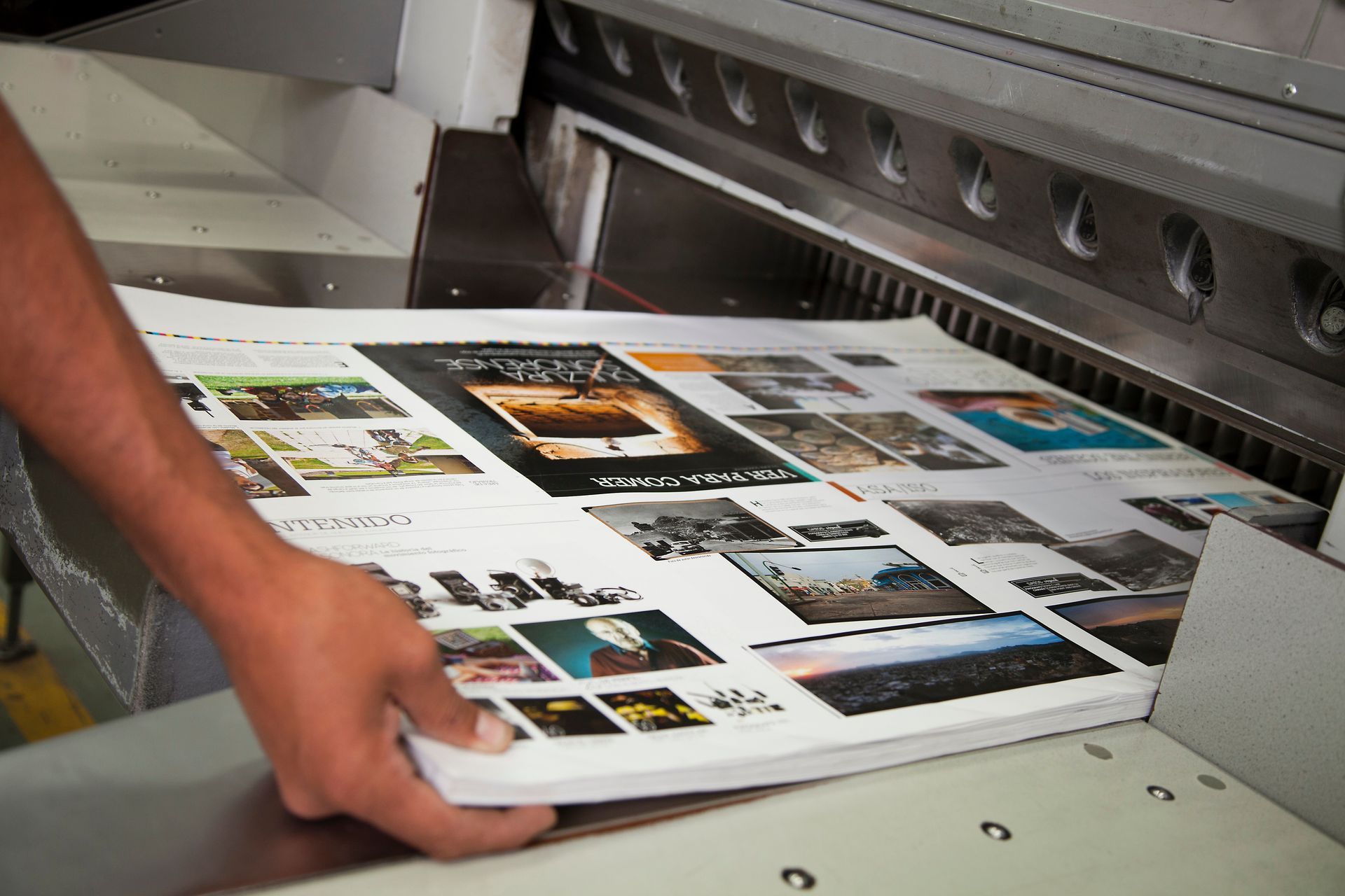 A person positions a large stack of printed pages under the blade of an industrial paper cutter in a print shop.