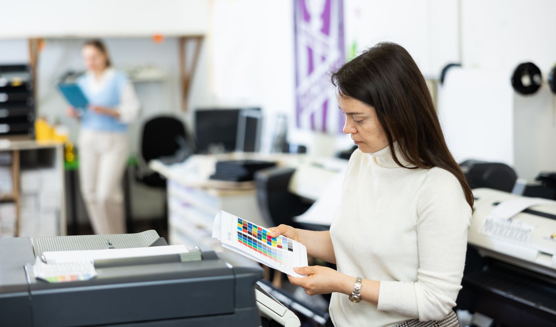 A person in a white turtleneck holds a color palette sample in an office with printing equipment.