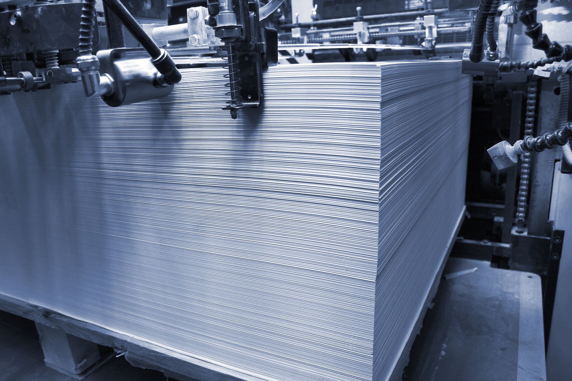 A large stack of printed paper sheets sits in an industrial printing press machine with a cool blue filter.