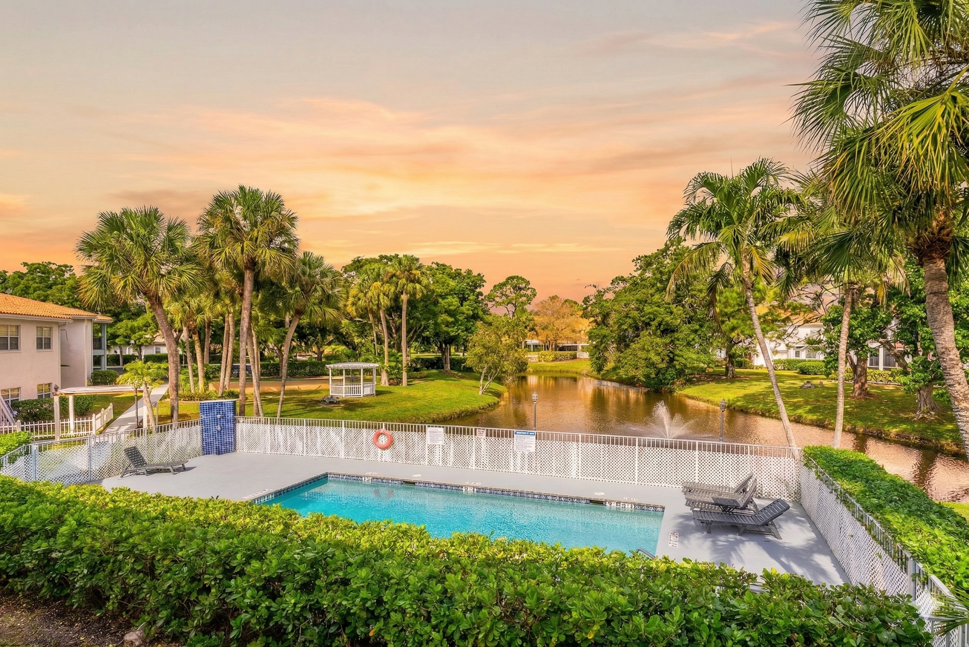A swimming pool enclosed by a white fence overlooks a small lake surrounded by palm trees under a sunset sky.