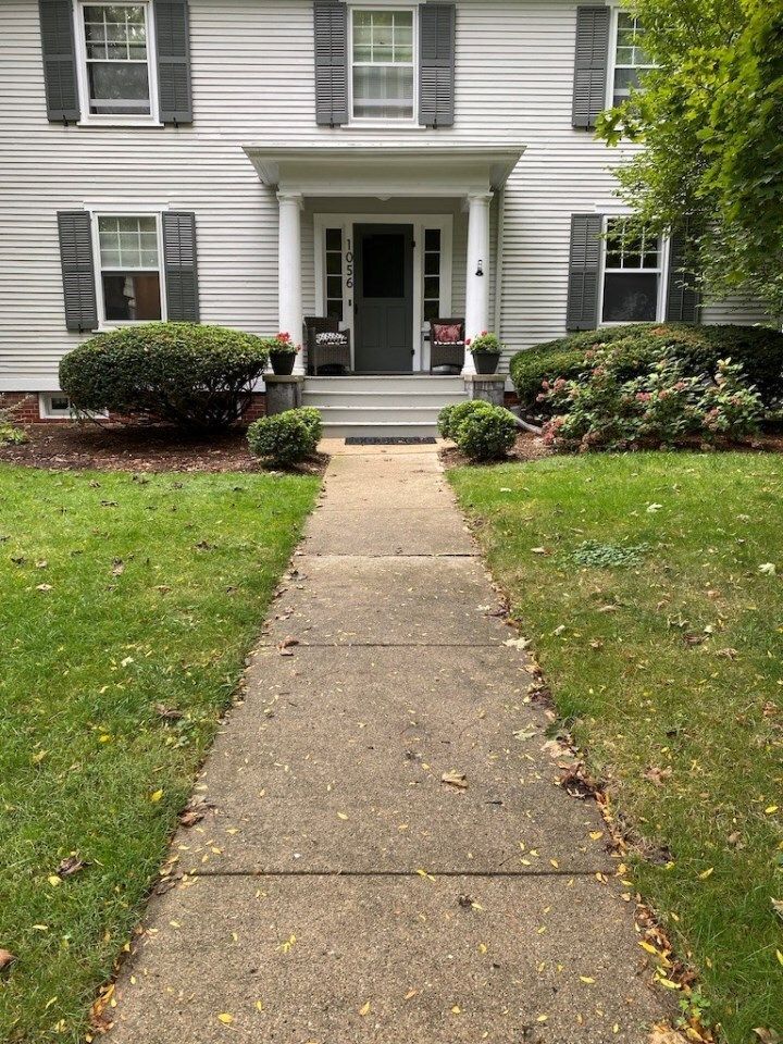 Front walkway to a white house with black shutters, green lawn, and a front porch with potted plants.