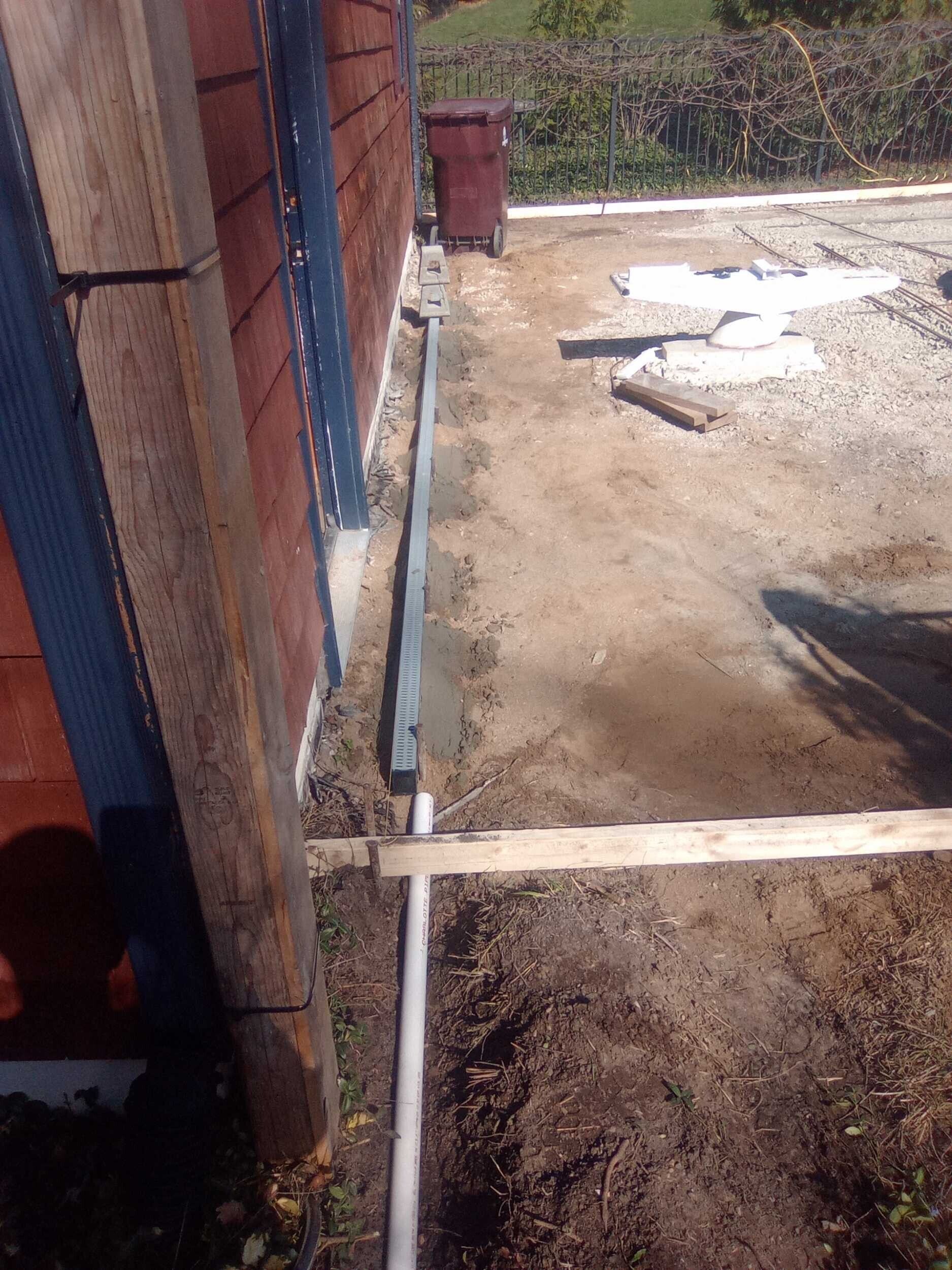 Narrow dirt path beside a building, with a wooden plank crossing and bright sunlight on the ground.