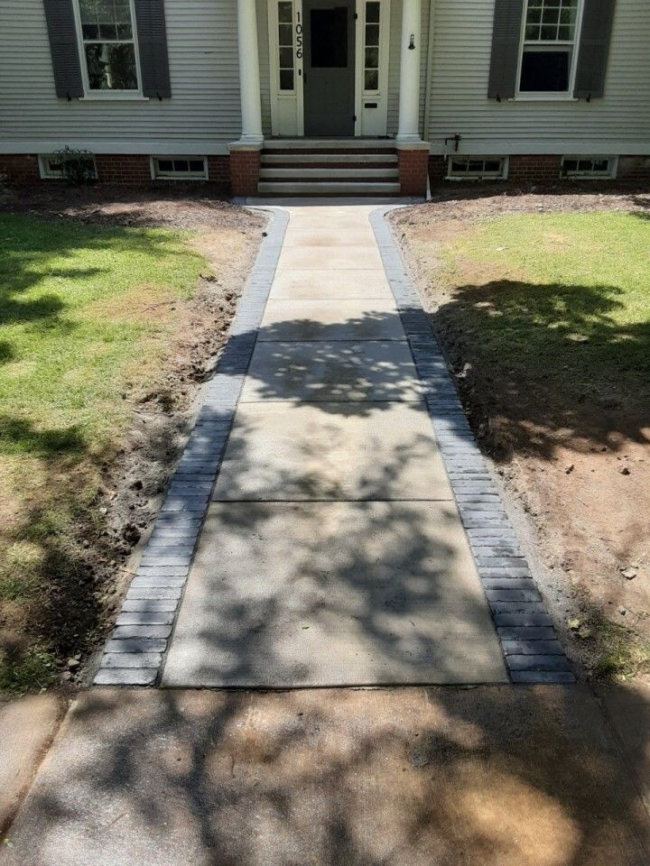 Concrete walkway leading to a house entrance with steps and brick edging, flanked by patchy grass.