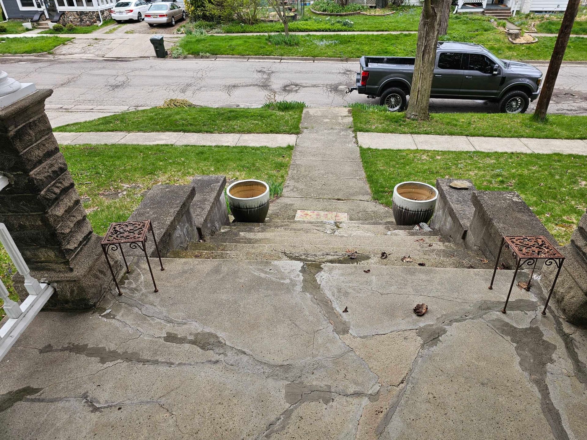 Front porch steps leading to a cracked concrete walkway, with potted plants and a street view beyond.