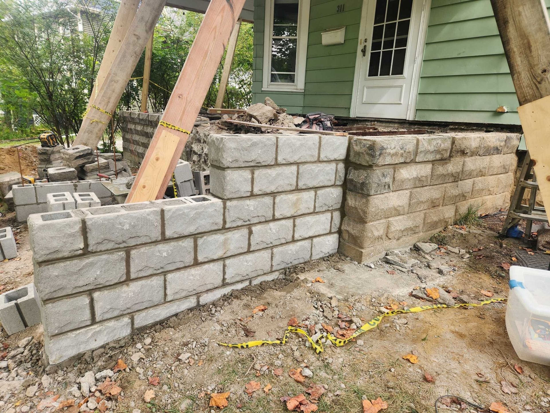 Partially built stone retaining wall beside a house with wooden supports and debris on the ground
