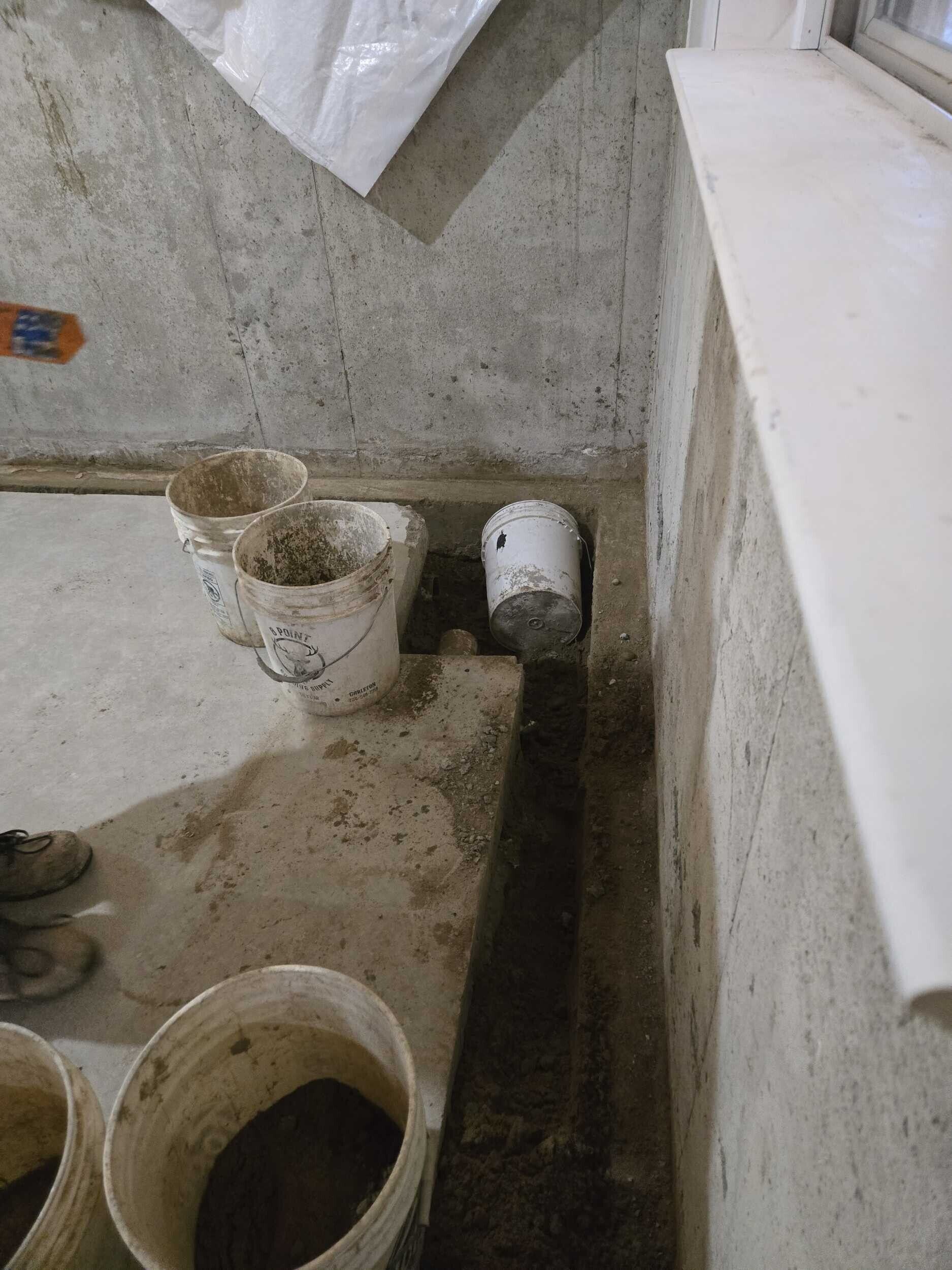 Dirty bathroom corner with a toilet bowl, bucket, and drain near a concrete wall and ledge