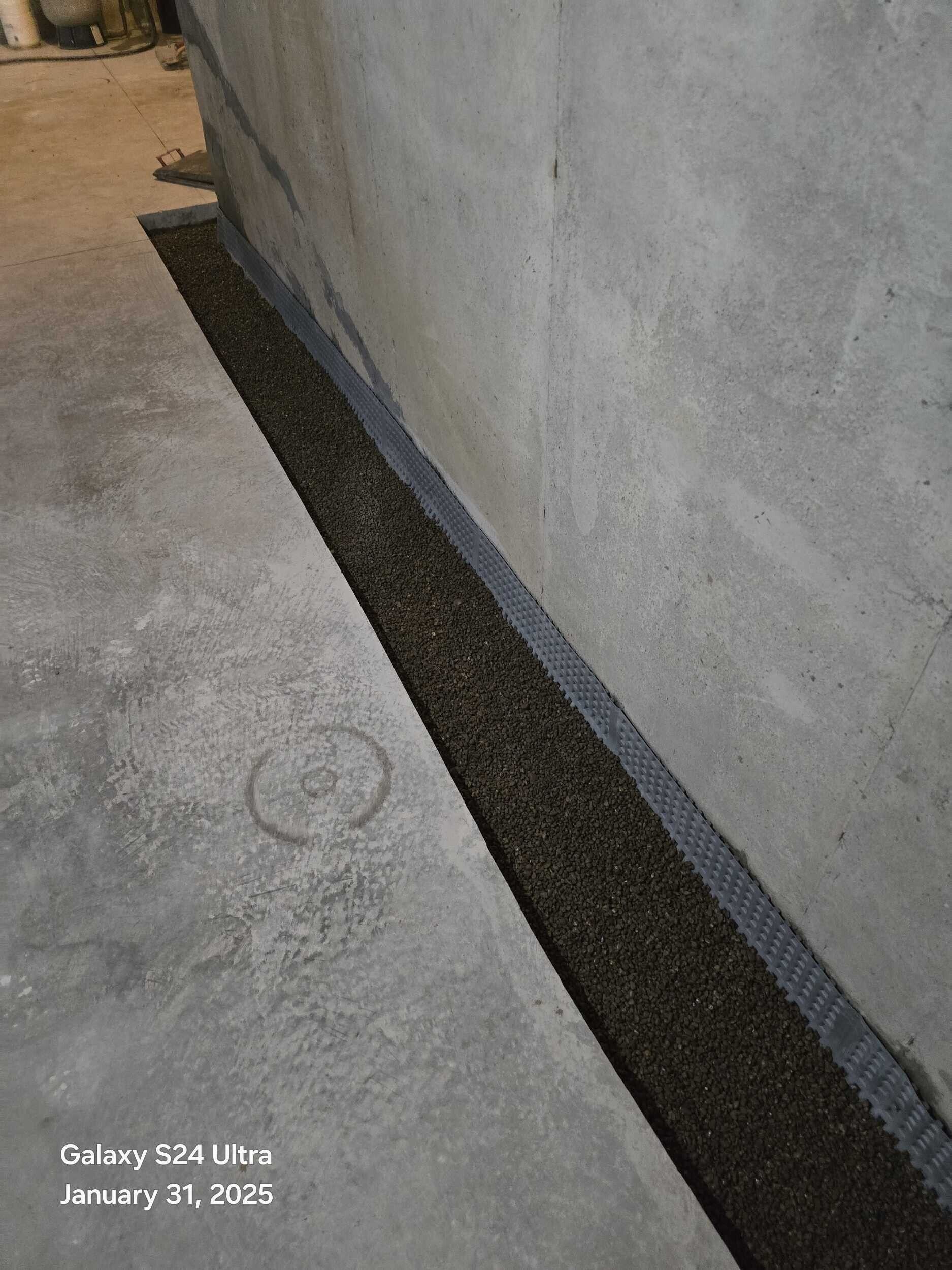 Narrow gravel drainage trench along the base of a white wall beside a concrete floor.