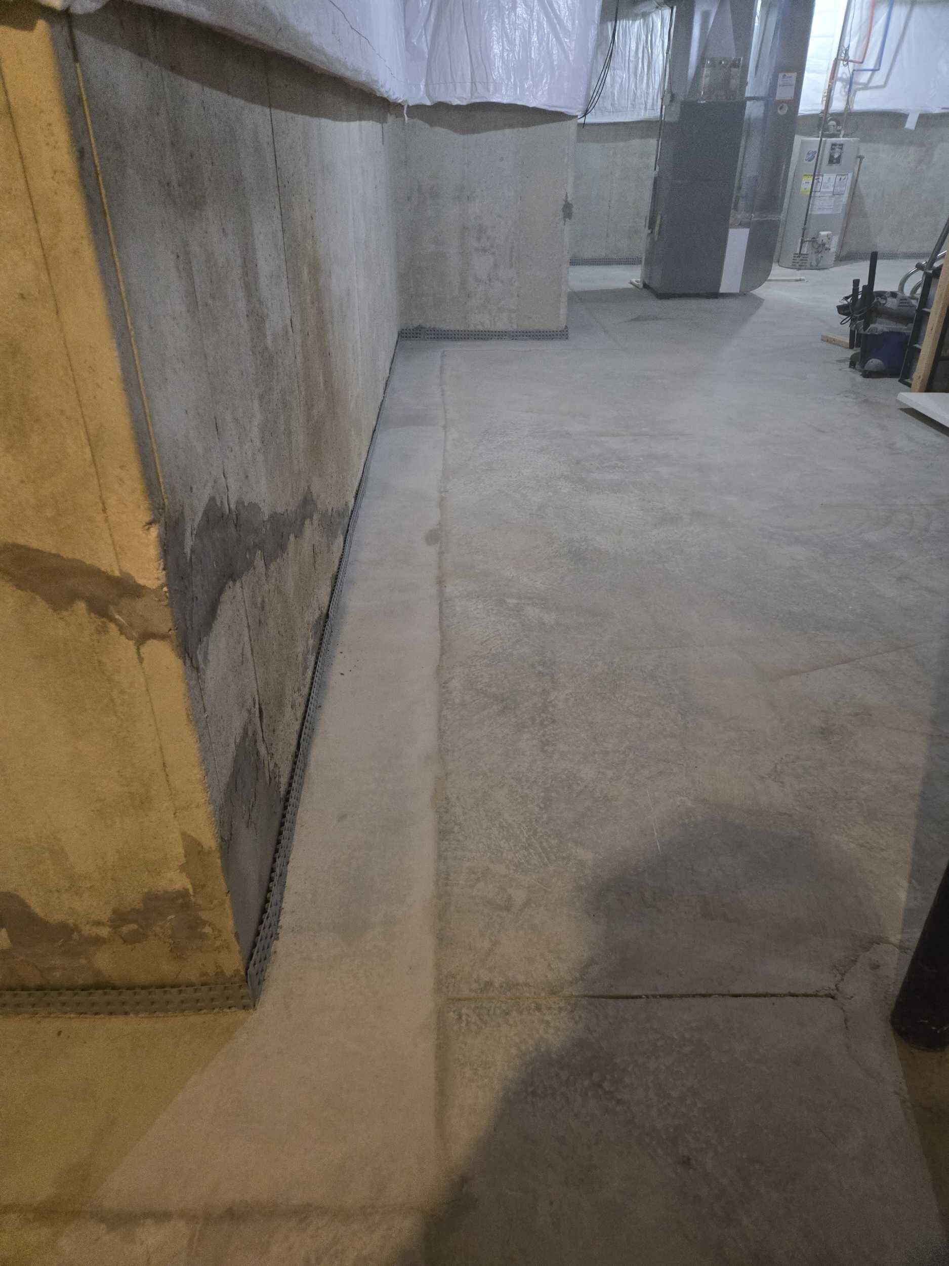 Concrete basement corridor with a stained wall and floor drain line, lit by overhead lights