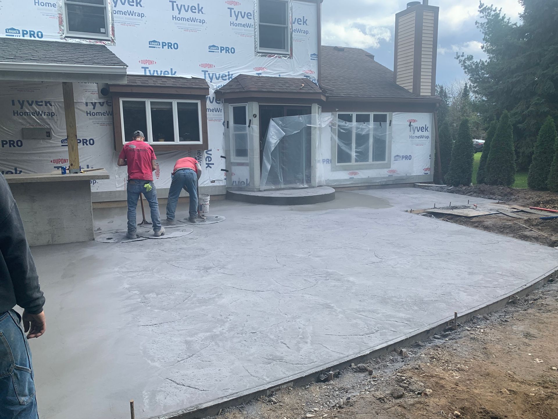 Two workers in red jackets stand on a newly poured concrete driveway beside a house under construction.