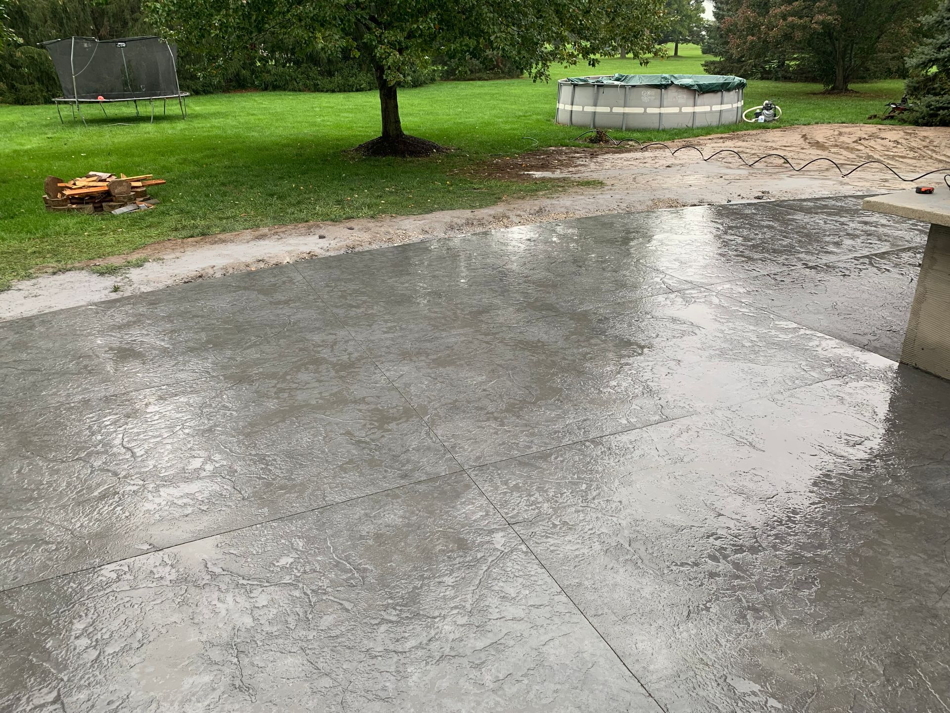 Wet concrete patio beside a grassy yard with trees and a trampoline