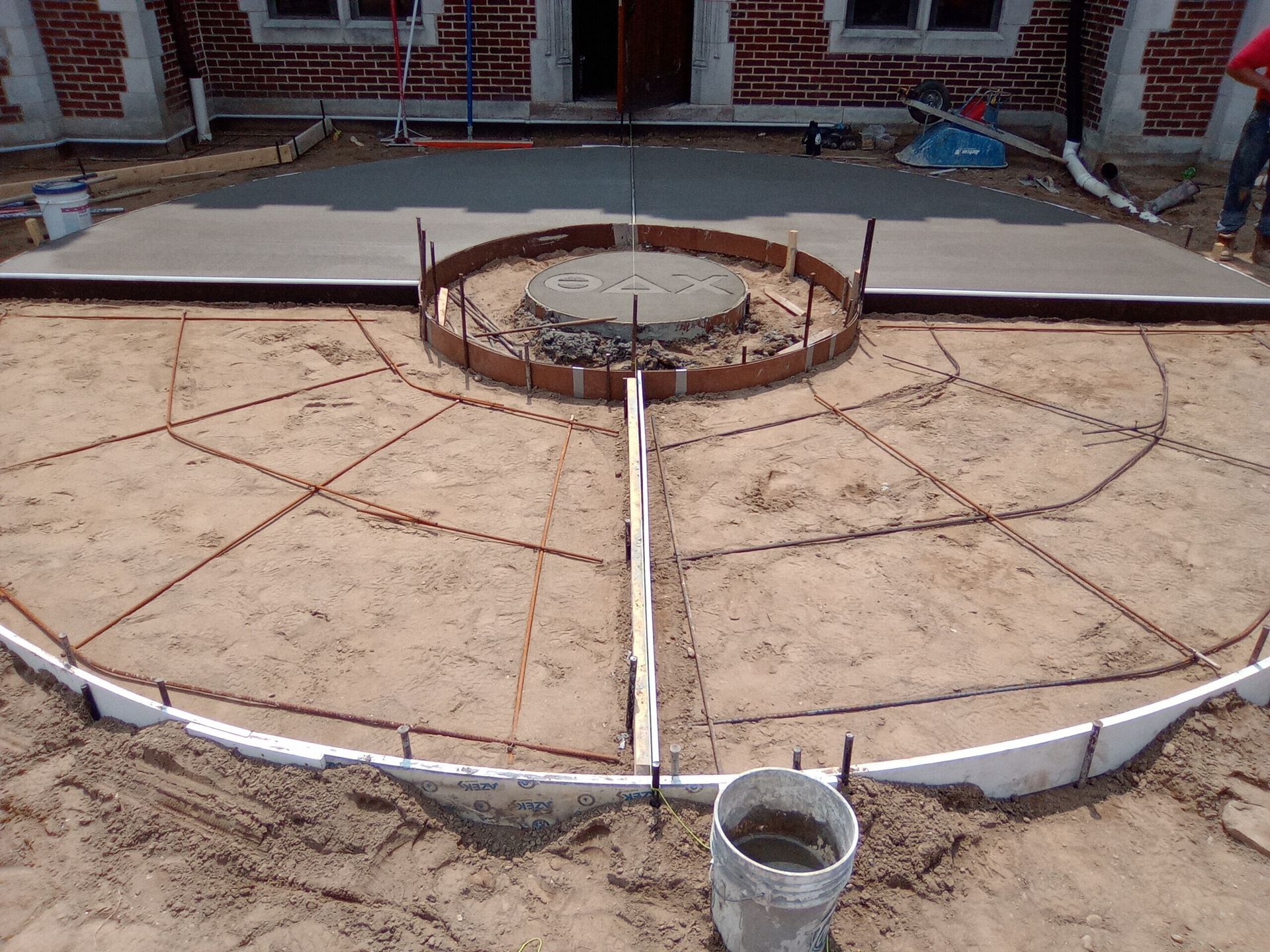 Construction site with a circular concrete foundation and central pit, marked by rebar and a bucket in front.