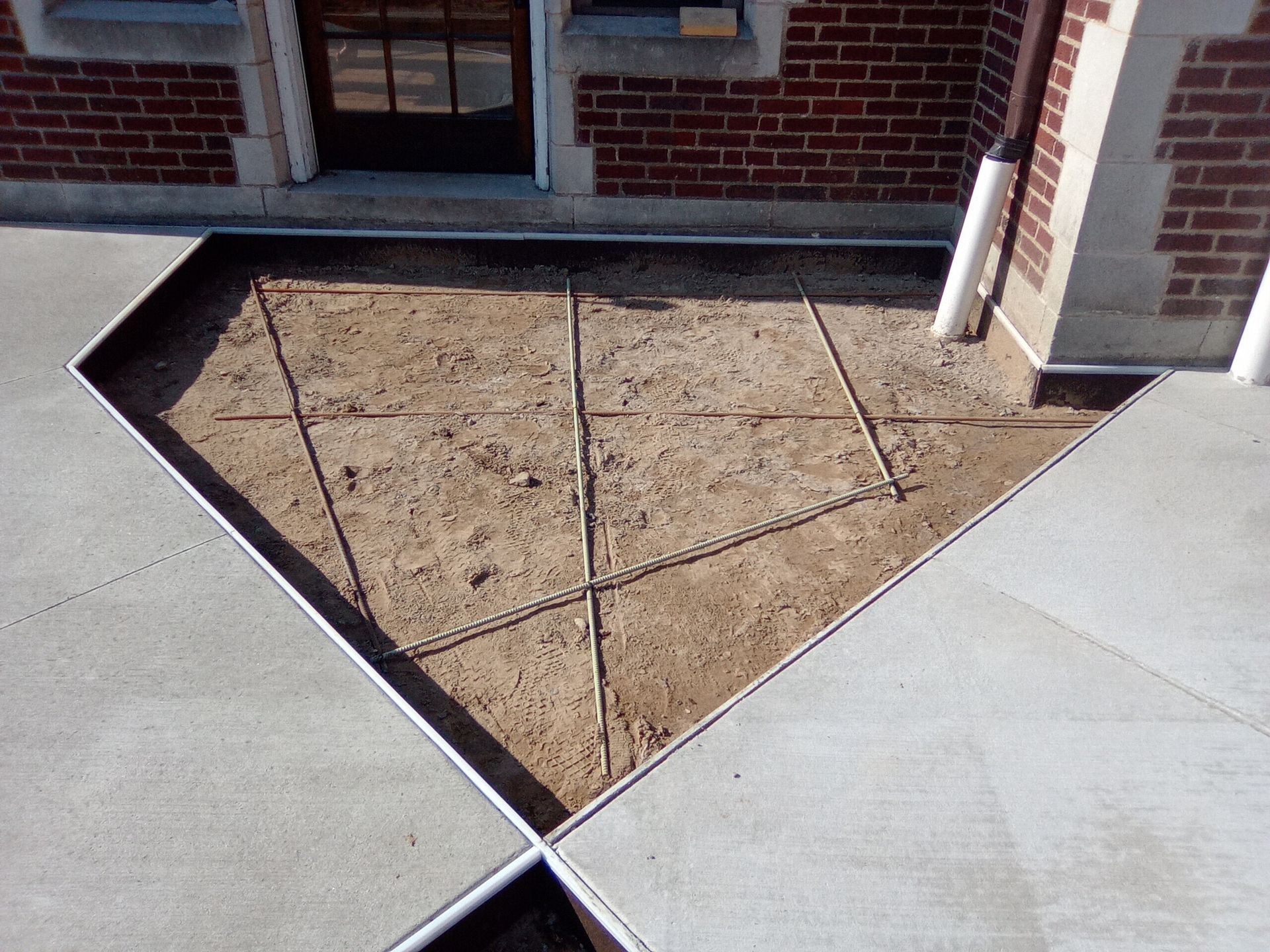 Triangular dirt planter bed bordered by white concrete beside a brick building and sidewalk