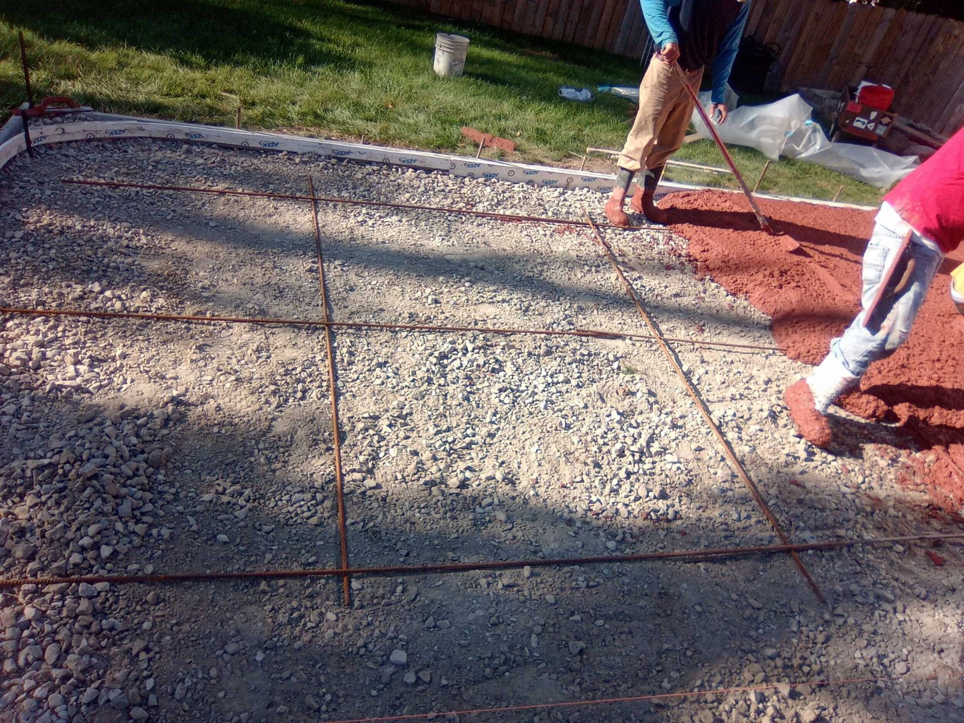 Two workers pouring and leveling red concrete on a driveway beside an existing gray slab.
