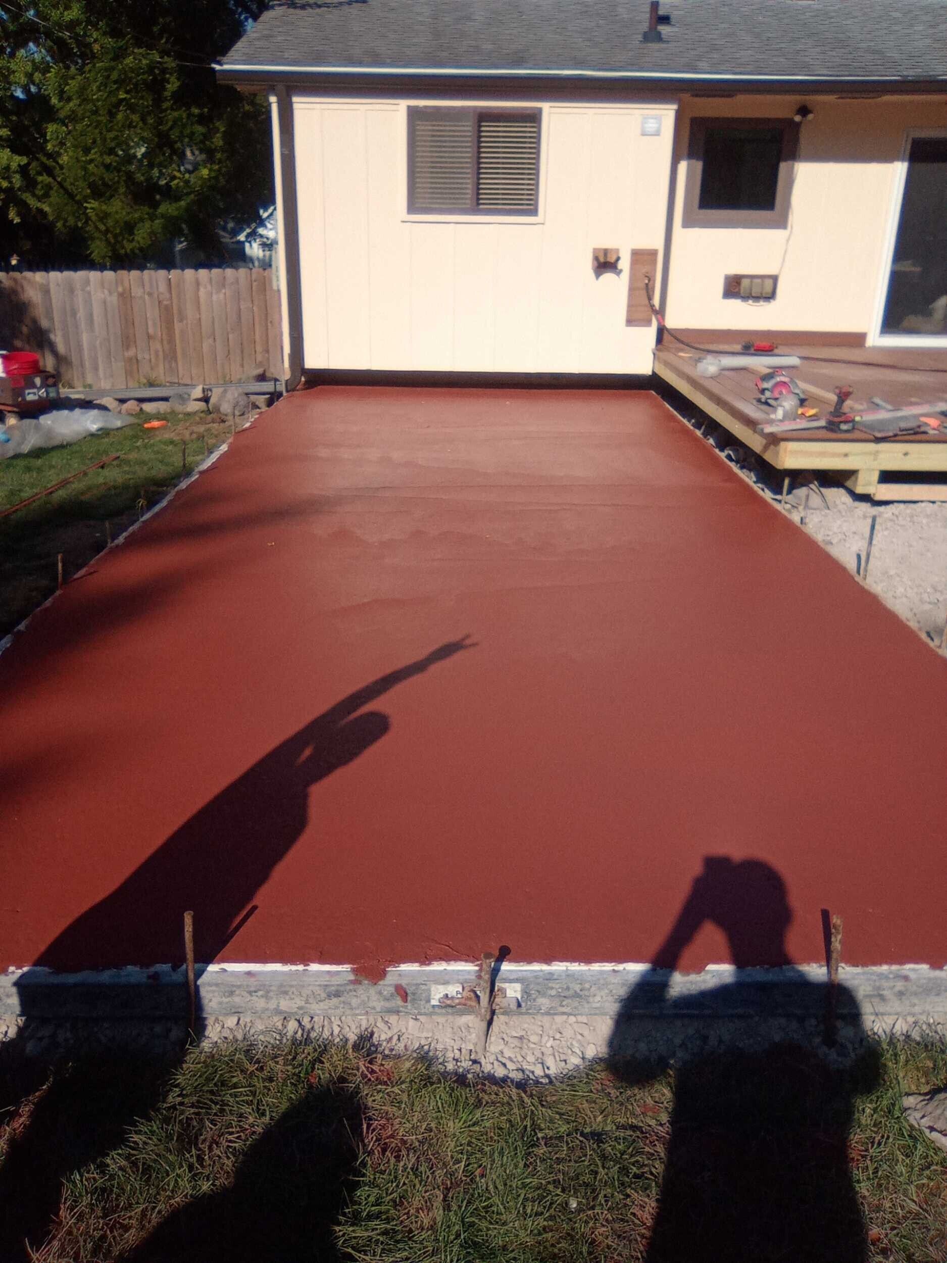 Freshly coated red concrete patio or driveway beside a house, with a worker’s shadow in the foreground