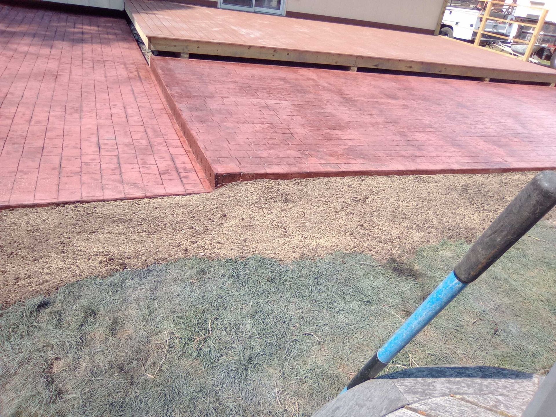 Close-up of a blue-handled rake over dry grass beside red brick paving and a raised walkway
