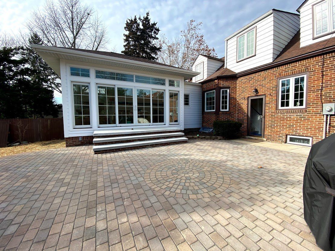 Patio with paver stones beside a white sunroom and brick house under a bright sky