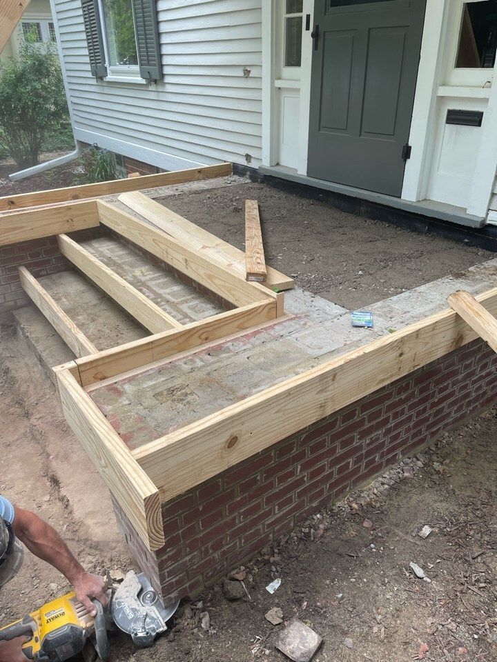 Partially built front porch steps and wooden frame beside a house, with a worker using a saw at lower left