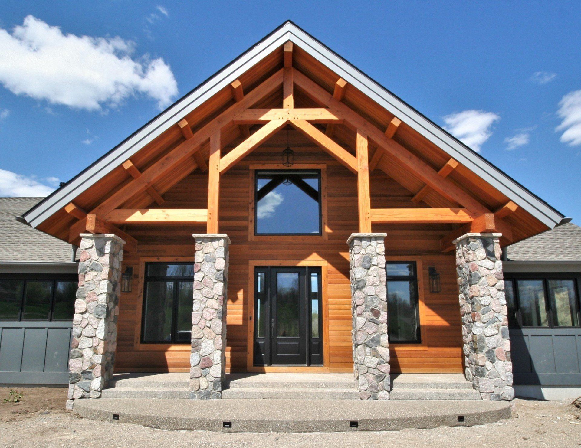 Modern log cabin-style house with stone pillars and a large peaked wooden entrance under a blue sky