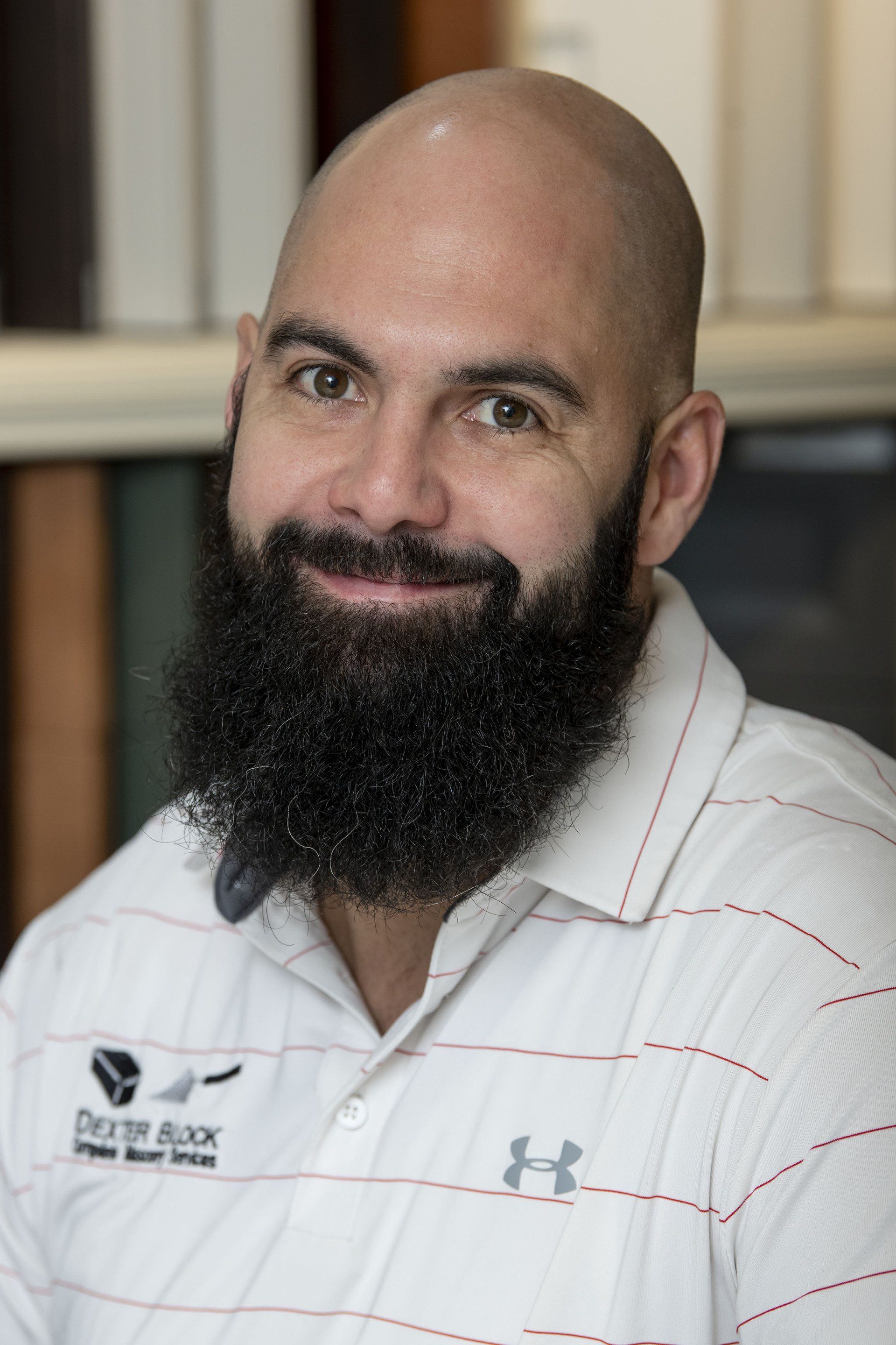 Smiling bald man with a long dark beard wearing a white striped polo shirt indoors