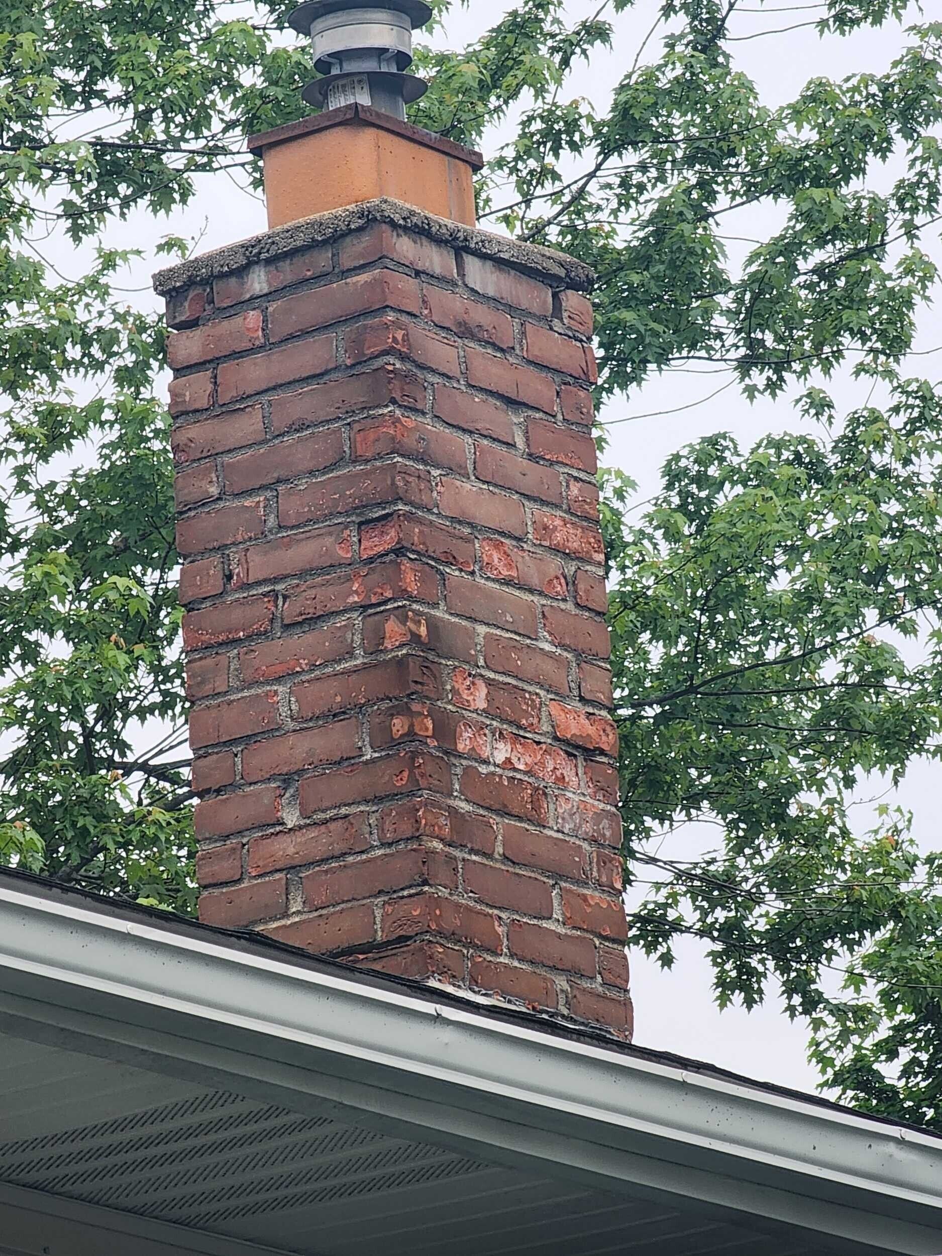 Brick chimney with a metal cap above a house roof, framed by leafy trees.