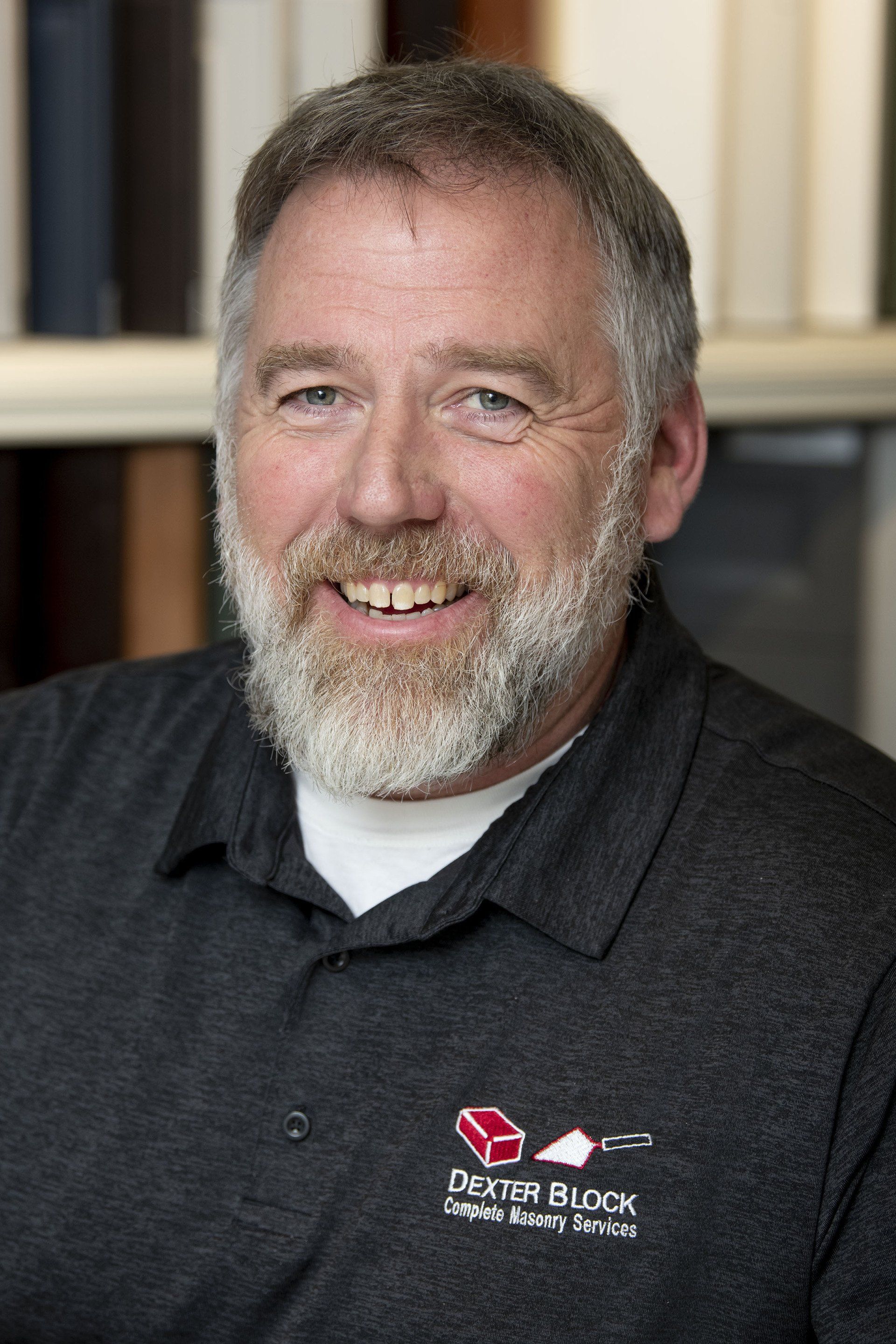 Smiling man in a dark polo shirt with a logo, seated indoors against a blurred bookshelf background